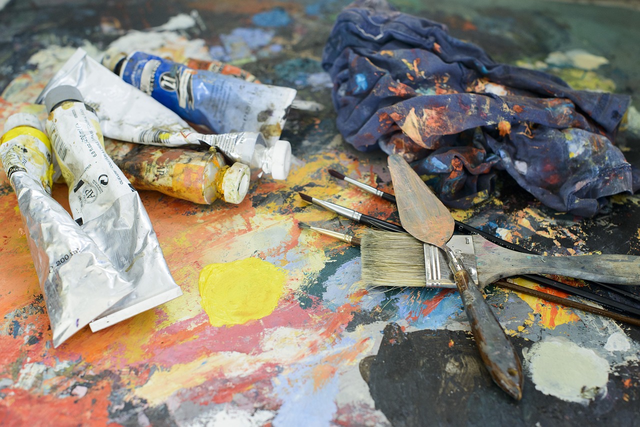 Tubes de peinture, pinceaux et spatules sur une table d’atelier couverte de couleurs.